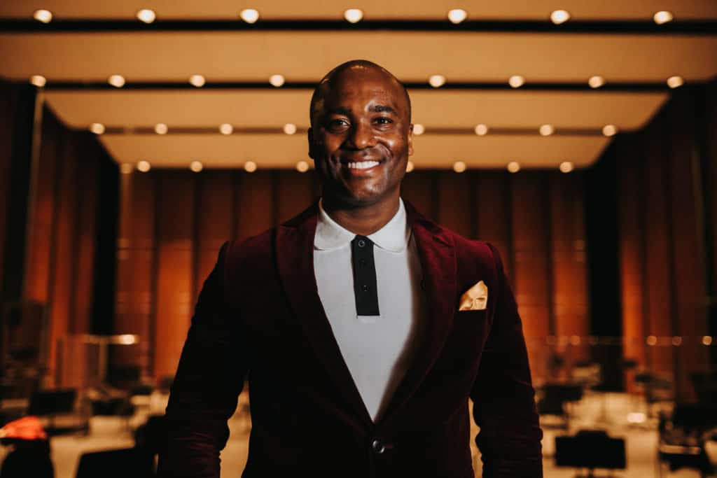 A smiling Black man in maroon blazer, with Dell Hall stage in the background