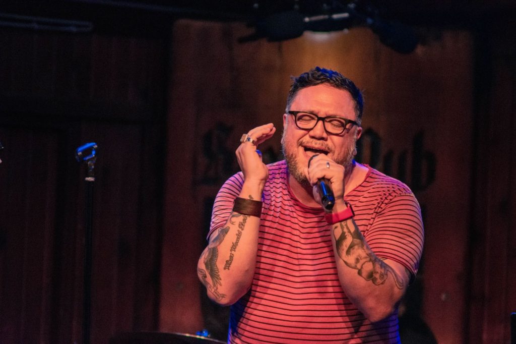 Nakia, a bespectacled, dark-haird man in a red striped shirt singing in a dark studio