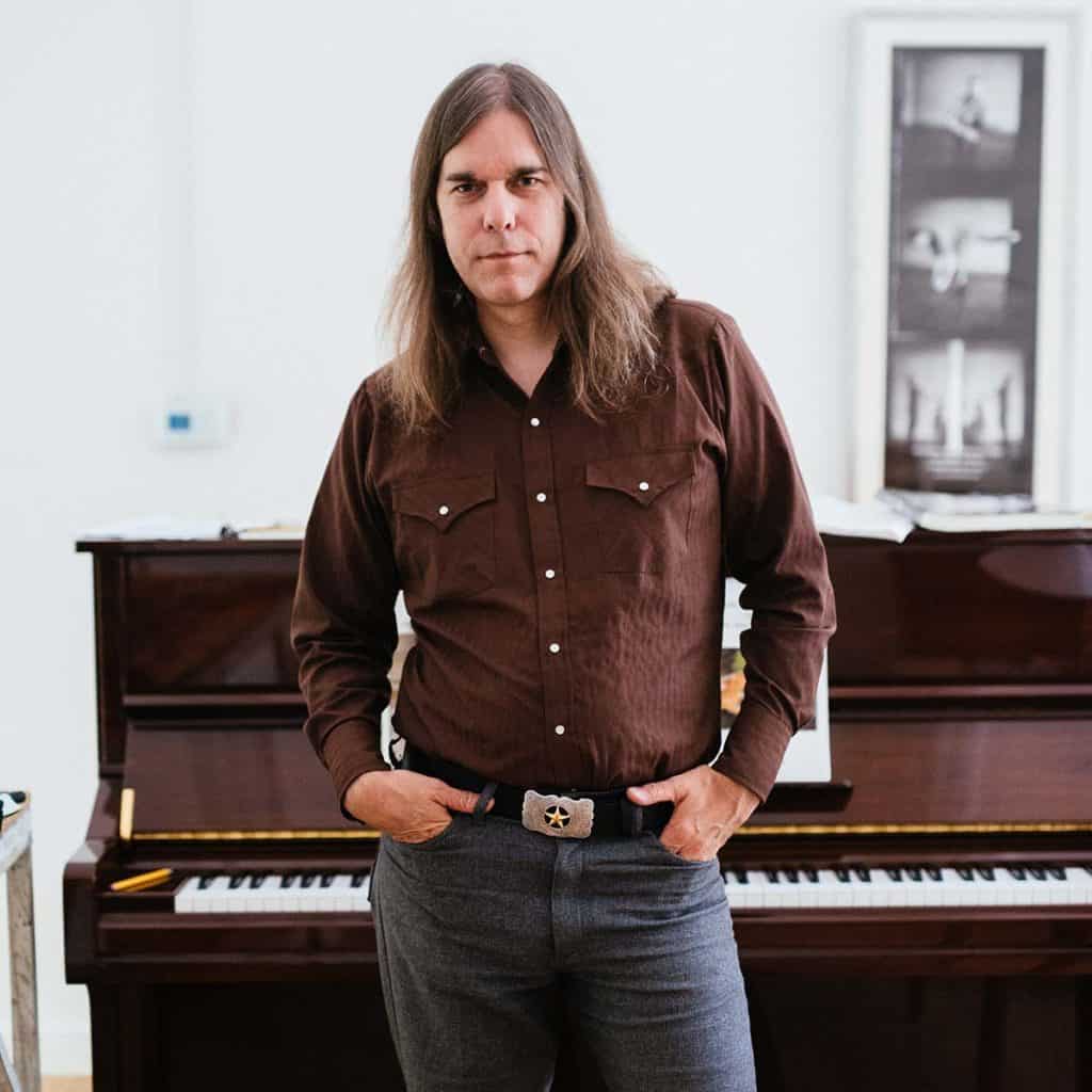 Graham Reynolds, a tall man with long brown hair stands in a brown pearl snap with his hands in his pockets in front of a brown studio upright piano.