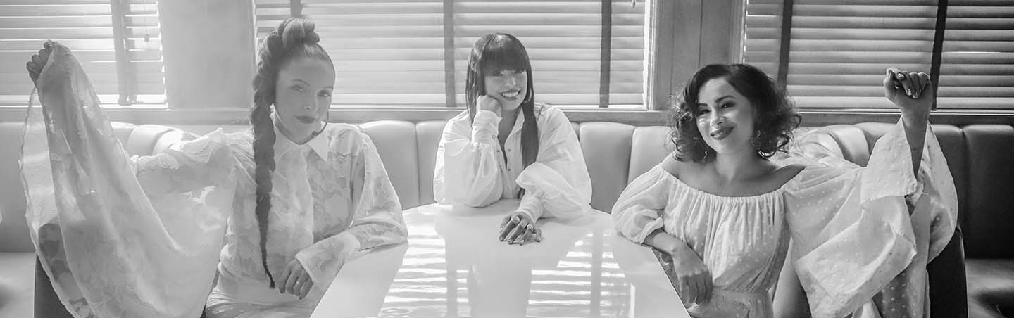 The ladies of Flor de Toloache pose all in white sitting in a diner booth in this black and white photo