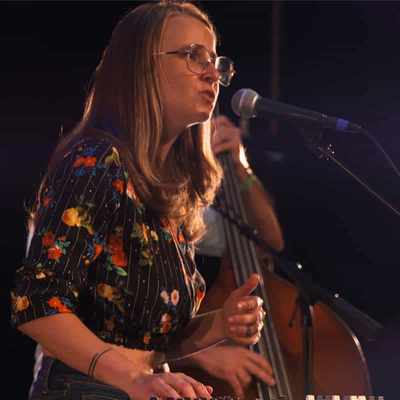 Grace, a white woman with long blonde hair and glasses, sings into a microphone backed by dark, moody stage lights