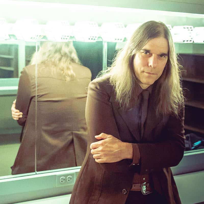 Graham, a white male with long brown hair, leans against the dressing room mirrors wearing a dark brown suit