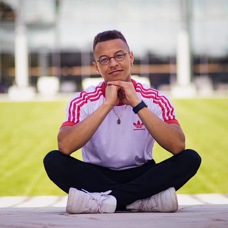 Donelvan, wearing glasses and a white and red t-shirt, sits cross-leged against a green field