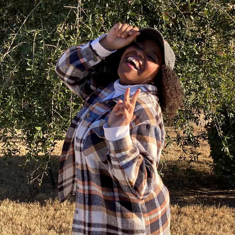 Jessica, poses with a big smile, a hat, and a peace sign in a tree-filled park